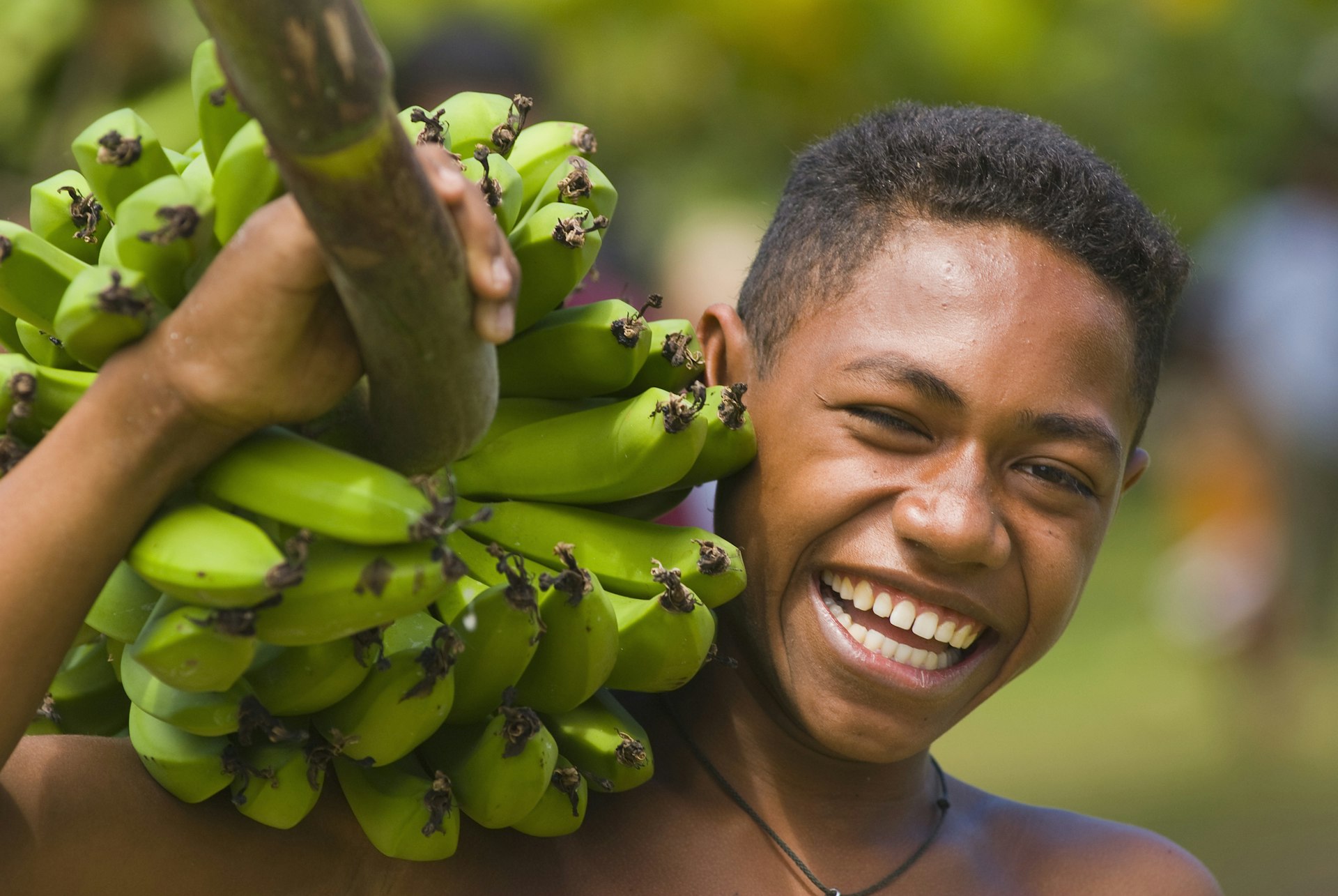A boy is carrying a bunch of bananas on his shoulder and laughing in Fiji